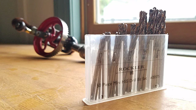 Box Of New Rockler Drill Bits With A Millers Falls No. 5 Egg Beater Antique Hand Drill In The Background On A Woodworking Workbench