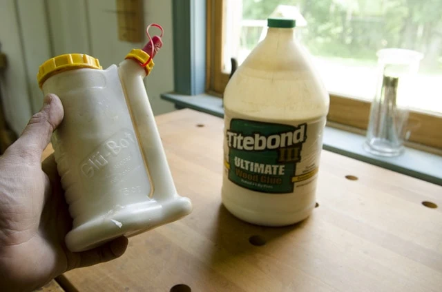 Glu Bot Glue Bottle Being Held With A Bottle Of Titebond 3 Wood Glue In The Background On A Woodworking Workbench