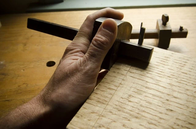 Woodworker Using A Wooden Cutting Gauge Marking Gauge On A Piec Of Figured Quartersawn White Oak Lumber Over A Roubo Woodworking Workbench With Other Marking Gauges In The Background
